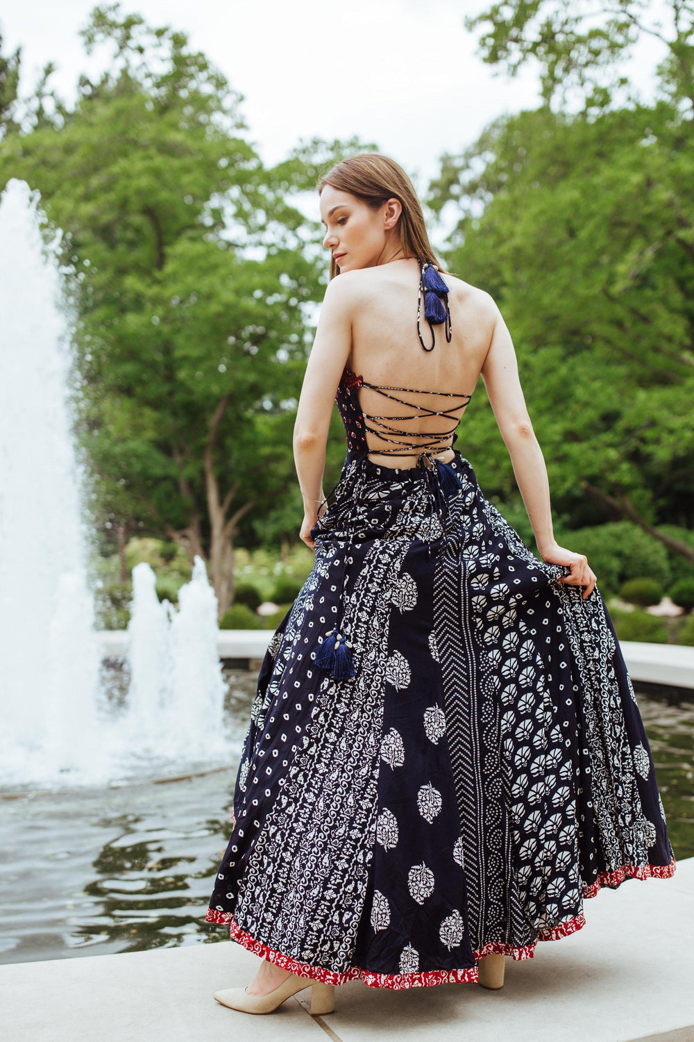 Woman in a navy printed cotton gown with an open, crisscross back and tassel details, standing by a fountain.