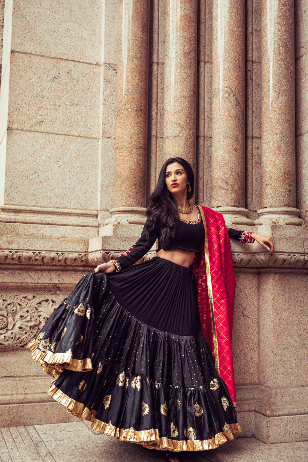 Woman in black silk full flare chaniya choli with gold butta, pearl embroidery, and red dupatta, posing against columns.