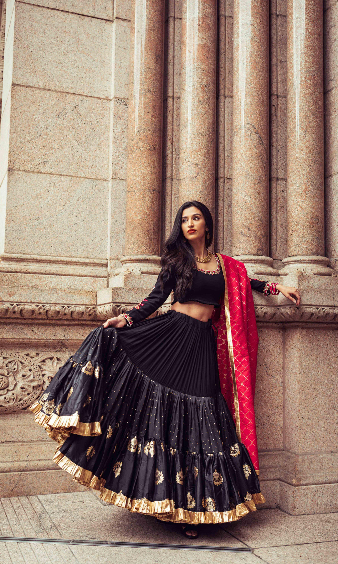 Woman in black silk full flare chaniya choli with gold butta, pearl embroidery, and red dupatta, posing against columns.