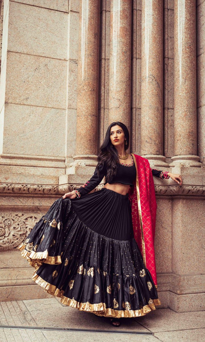 Woman in black silk full flare chaniya choli with gold butta, pearl embroidery, and red dupatta, posing against columns.