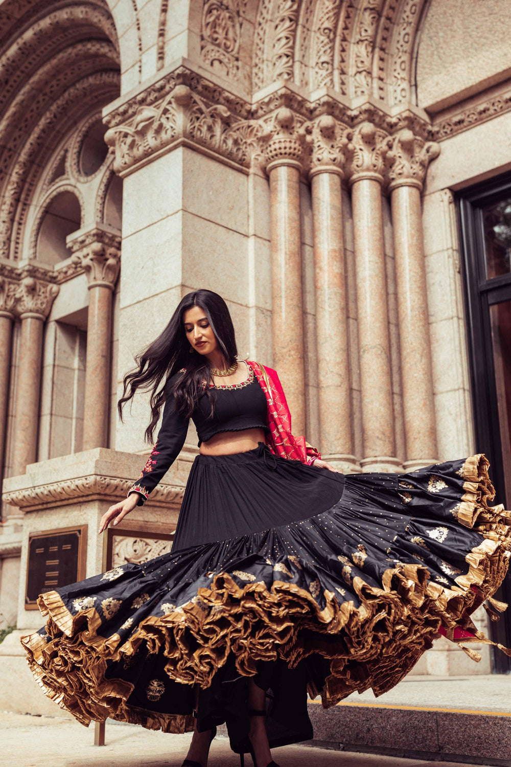 Woman in a black silk full-flare chaniya choli with gold butta, pearl embroidery, and a red dupatta, dramatically twirling.