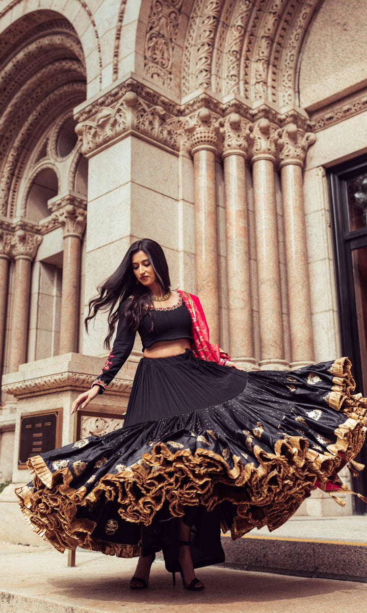 Woman in a black silk full-flare chaniya choli with gold butta, pearl embroidery, and a red dupatta, dramatically twirling.
