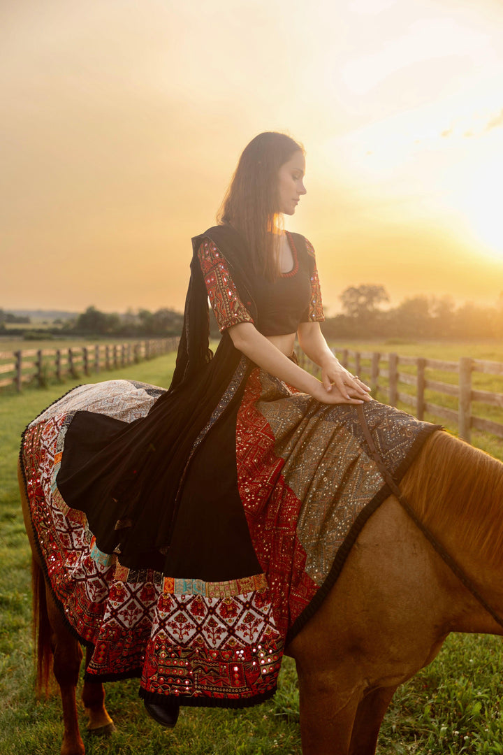 Black silk lehenga choli with multi-colored side panel and embroidered border, worn by a woman on a horse at sunset.