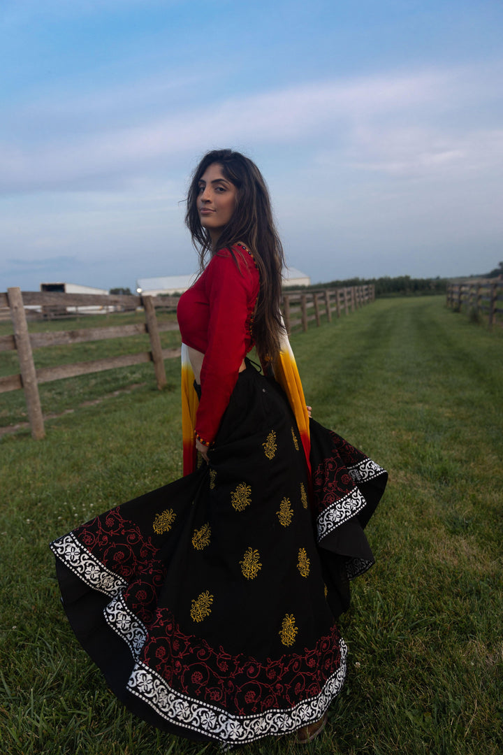 Woman in a dramatic twirl wearing a black cotton lehenga with yellow-gold and red print, red long-sleeve blouse, and shaded dupatta.