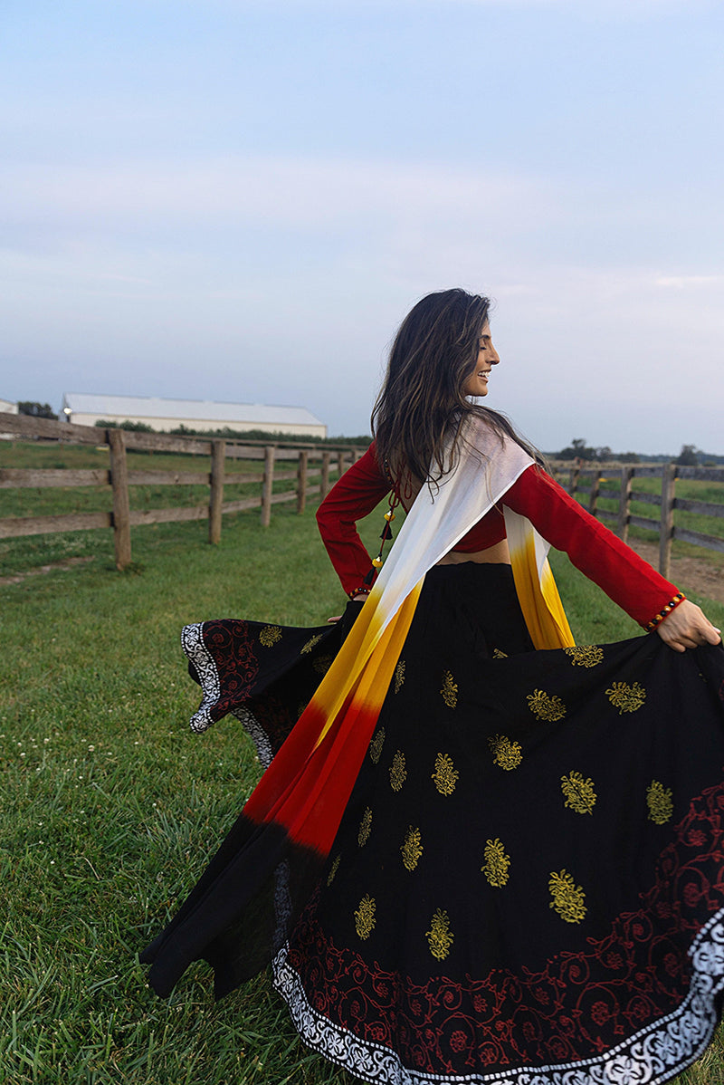 Woman in a black, red, and yellow cotton Chaniya Choli set with a long-sleeve blouse, dramatically twirling in a field.
