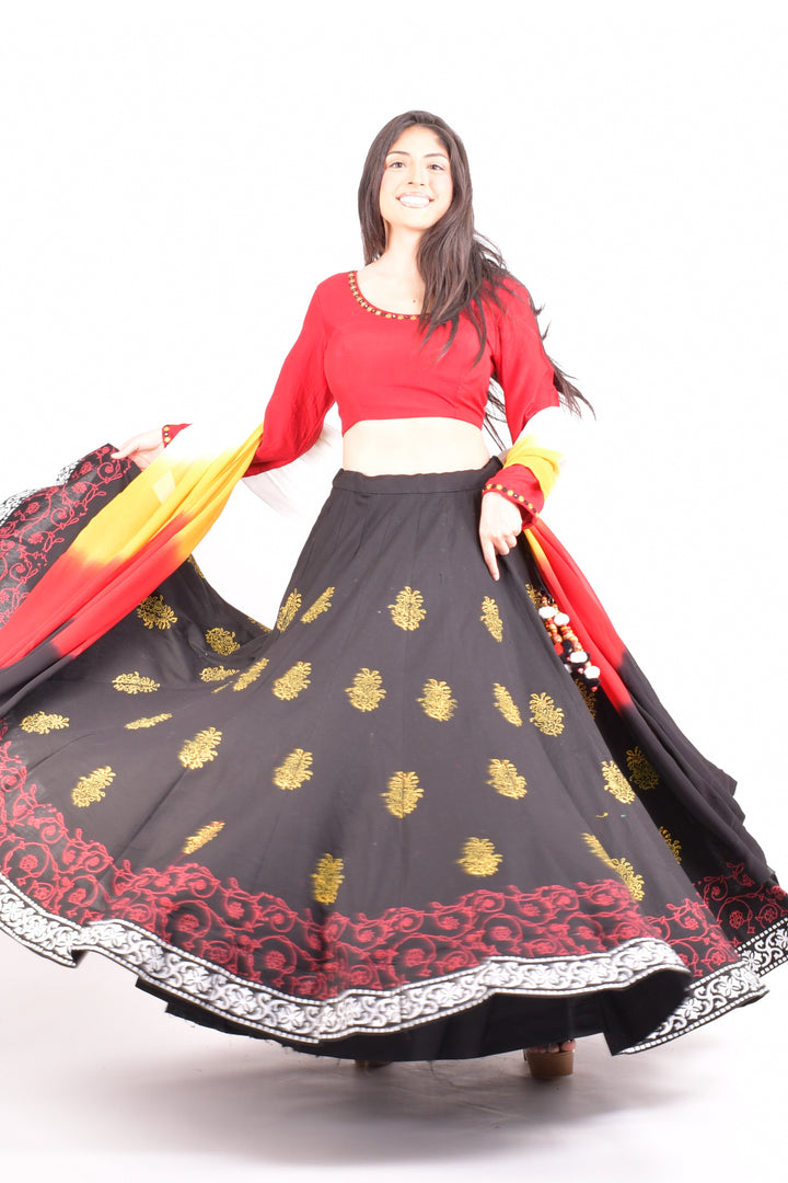 Woman in black cotton lehenga with gold print, red long-sleeve blouse, and shaded red-orange-yellow dupatta, dramatic twirl.