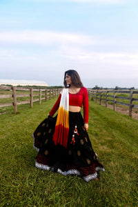 Woman in a black cotton lehenga with gold print, red long-sleeve blouse, and shaded white, orange, red dupatta, twirling outdoors.