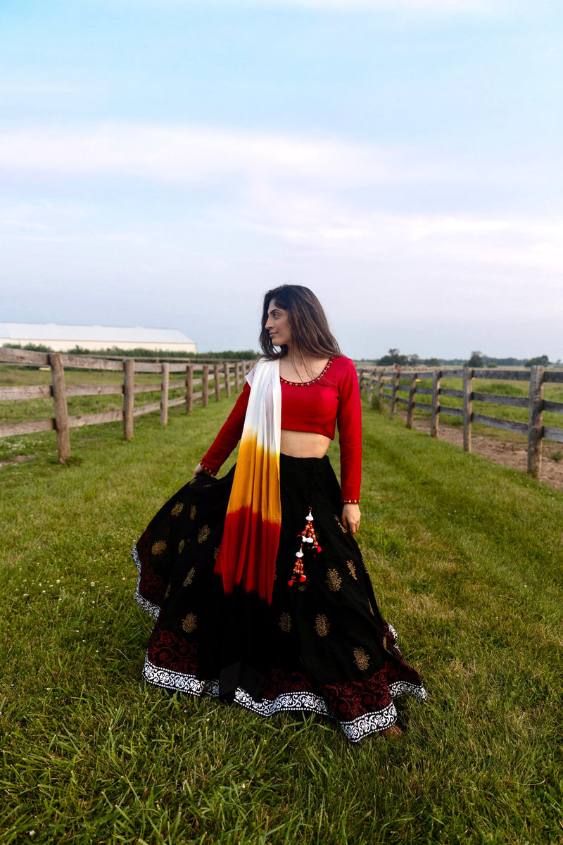 Woman in a black cotton lehenga with gold print, red long-sleeve blouse, and shaded white, orange, red dupatta, twirling outdoors.