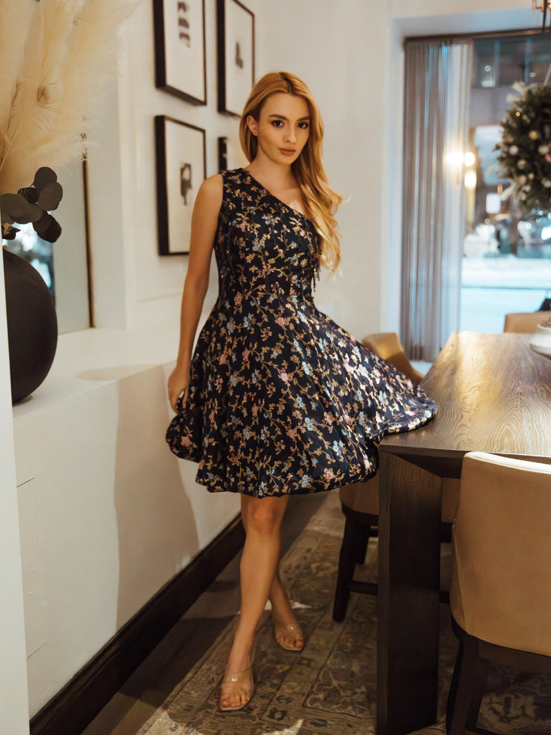 Model in a navy jacquard silk one-shoulder short dress with floral embroidery and a fit-and-flare silhouette.