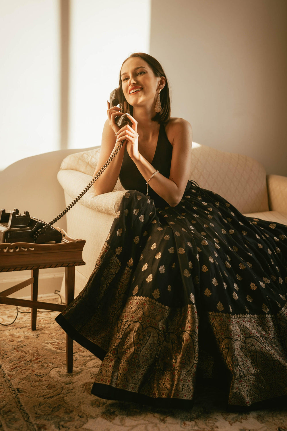 Woman in a black silk gown with a gold Banarasi border, V-neck, and open back, holding a vintage phone.