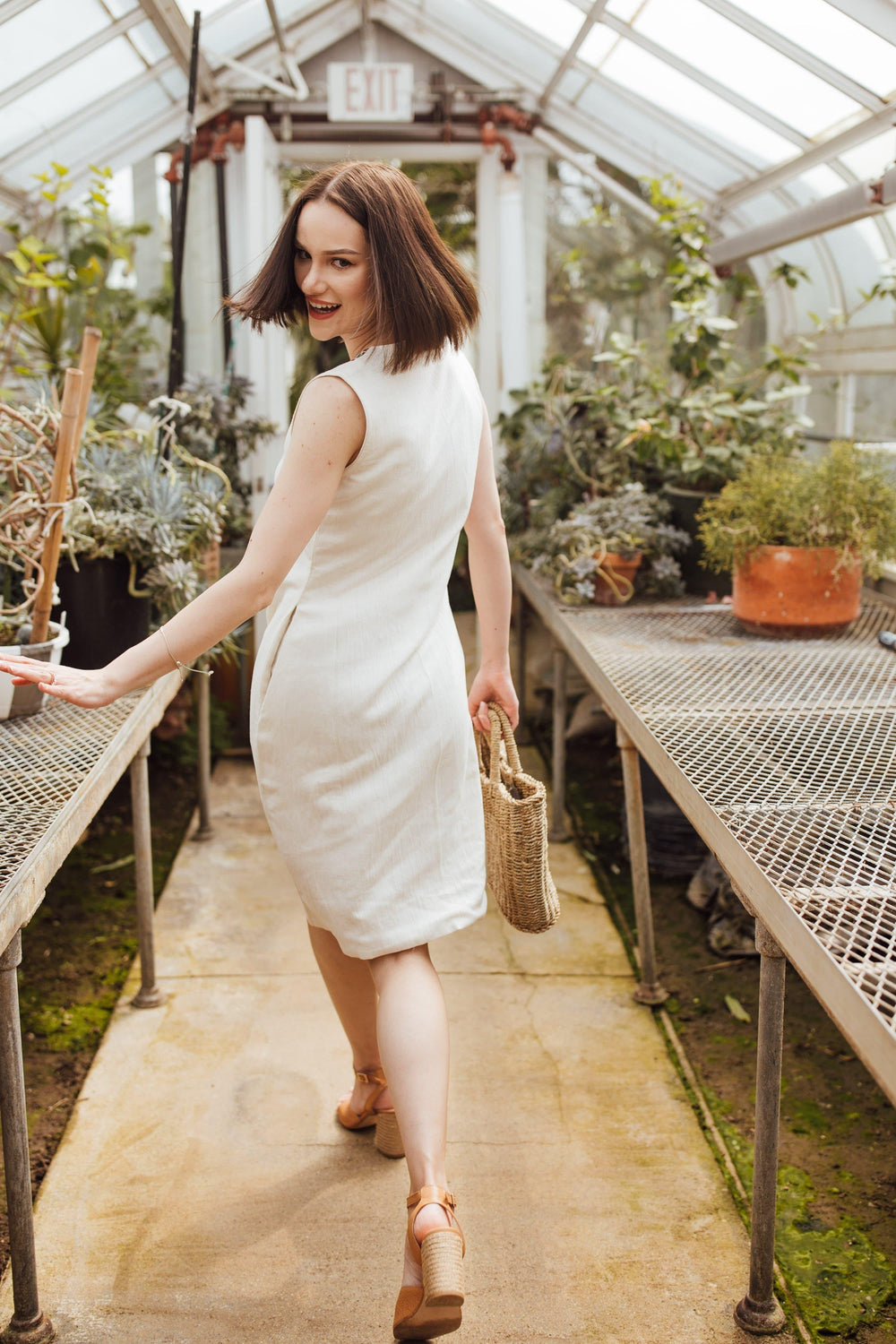 Woman in an off-white sleeveless cotton midi dress with a block-printed neckline, holding a straw bag.
