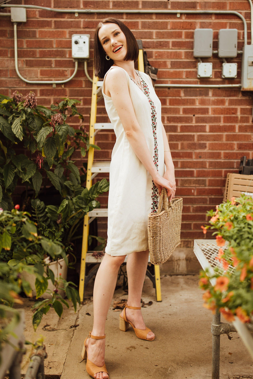 Off-white sleeveless cotton midi dress with a block-printed neckline, worn by a woman holding a straw bag.