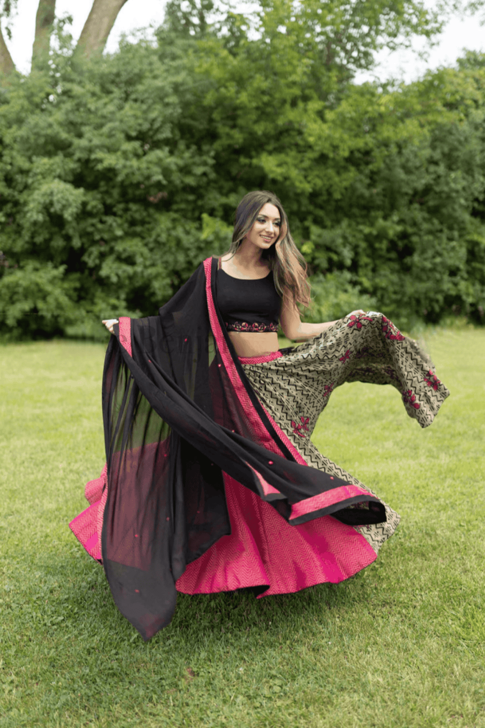 Woman in embroidered multicolor cotton-silk Chaniya Choli set, black blouse, fuchsia skirt, and black dupatta, twirling outdoors.