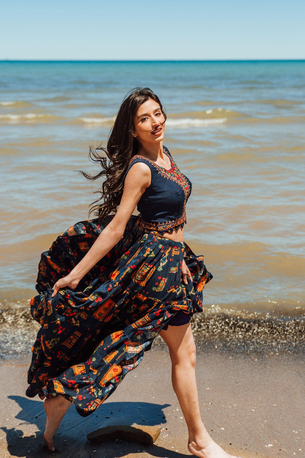 Embroidered navy high-low skirt set with a crop top and printed skirt, worn by a model on a beach.