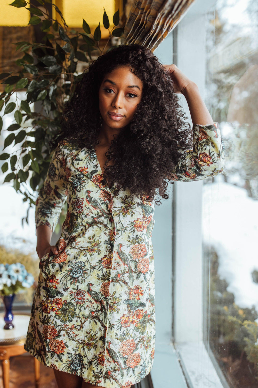 Woman in a floral eyelet embroidered cotton-silk blazer dress with long sleeves, standing by a window.
