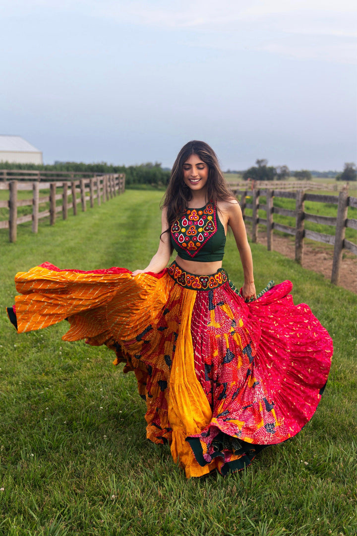Woman in a full flare multicolor Chaniya Choli with a green embroidered top and pink dupatta, twirling in a field.