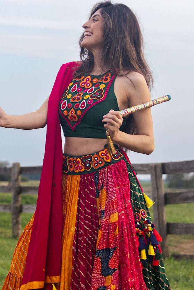 Woman in a full flare multicolor cotton Chaniya Choli, green embroidered blouse, pink dupatta, holding dandiya sticks.