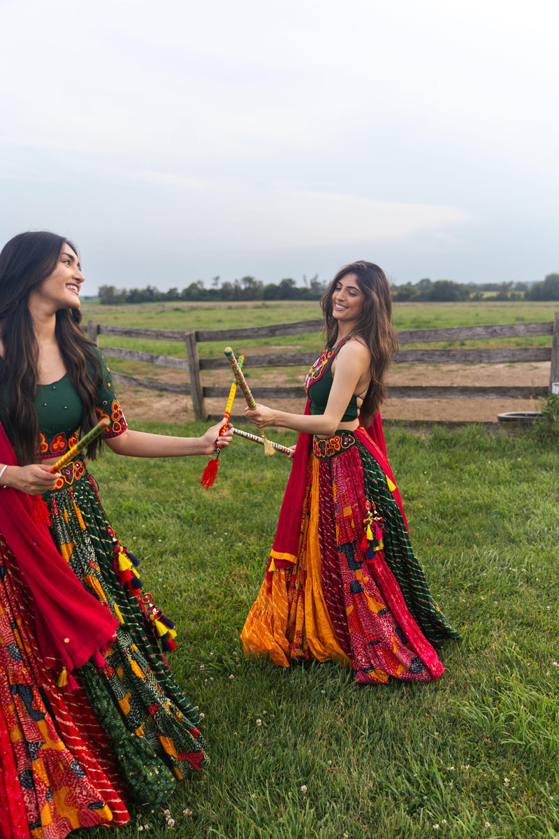 Two women in full flare multicolor cotton Chaniya Choli sets with green blouses and pink dupattas, holding dandiya sticks, performing Garba outdoors.