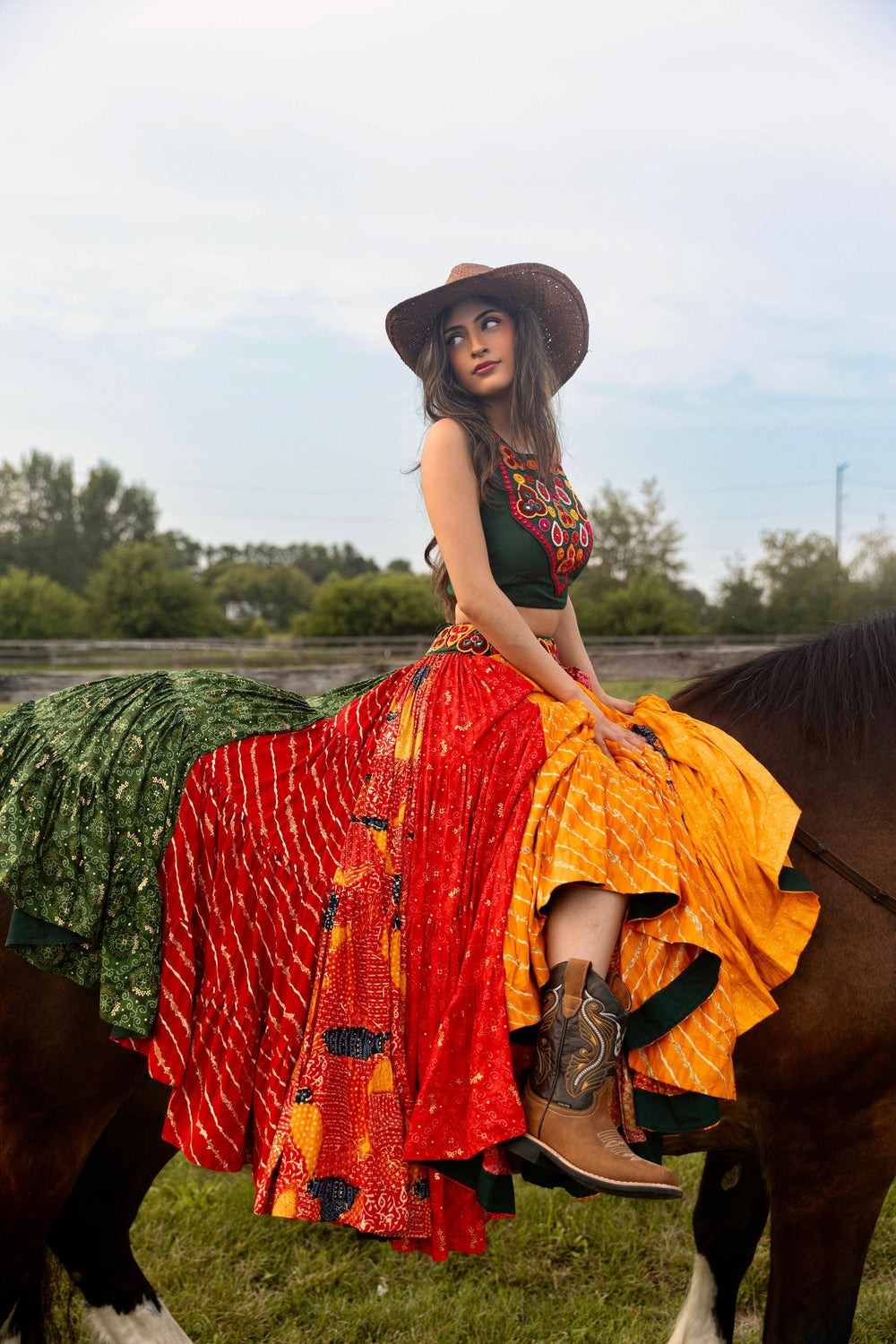 Woman in a full flare multicolor Chaniya Choli, green embroidered top, and cowboy hat, sitting on a horse.