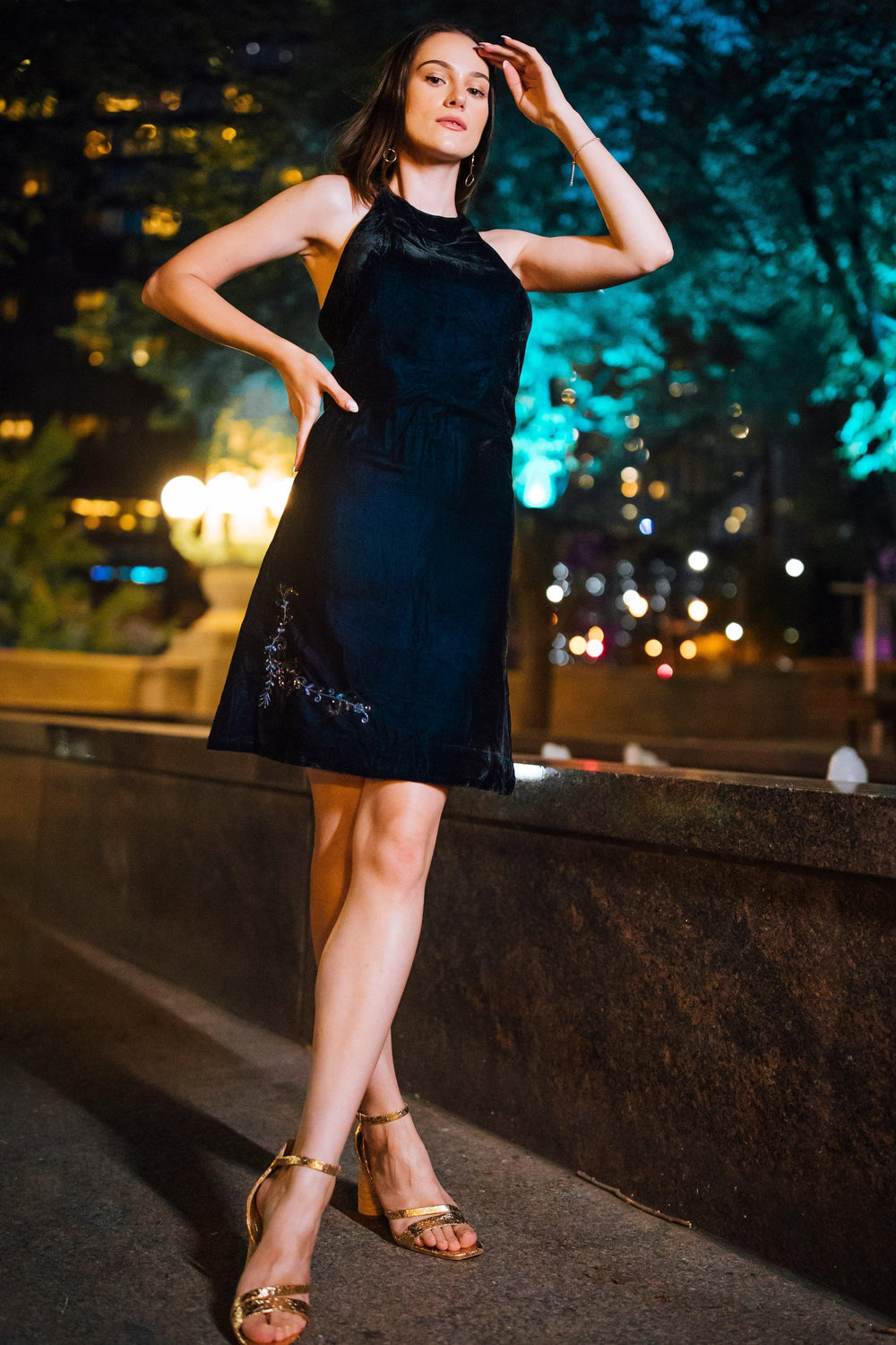 Model in a black velvet halter mini dress with hand embroidery and gold heels.