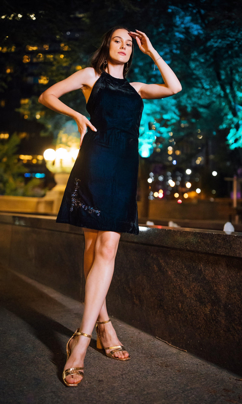 Model in a black velvet halter mini dress with hand embroidery and gold heels.