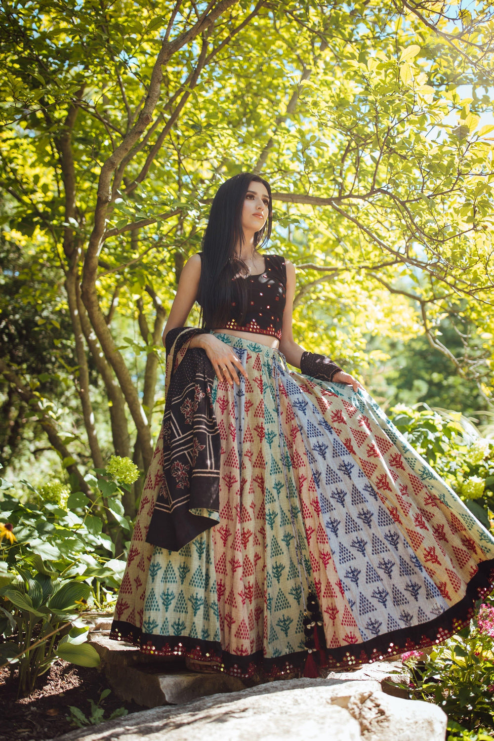 Woman in a black, sleeveless, mirror-work embroidered cotton blouse and a full-flare, hand-block printed lehenga with a mirror-work border, holding a black dupatta, posing outdoors.