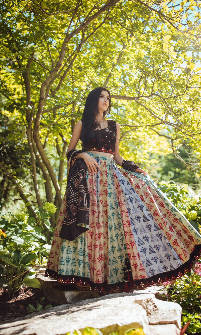 Woman in a black, sleeveless, mirror-work embroidered cotton blouse and a full-flare, hand-block printed lehenga with a mirror-work border, holding a black dupatta, posing outdoors.
