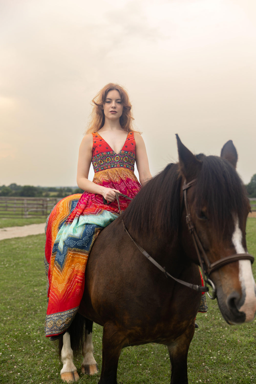 Woman in a vibrant silk bandhani print long dress with an embroidered bodice, riding a brown horse.