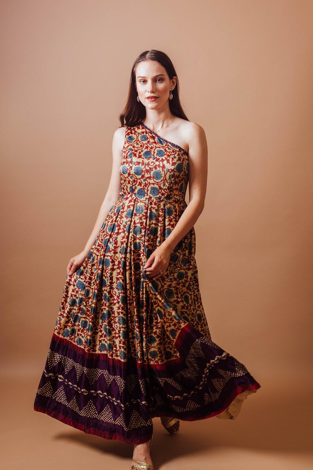 Woman in a flowy Gaji silk one-shoulder dress with intricate blue and red ajrakh print and a bold hem.