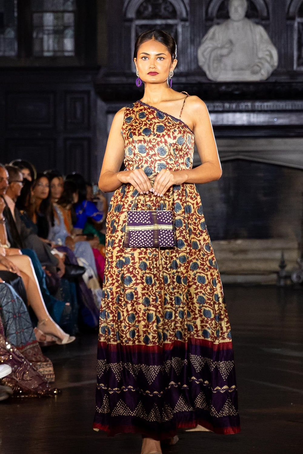 Model in a flowy Gaji silk one-shoulder dress with all-over ajrakh print and contrasting hem, holding a small purple bag.