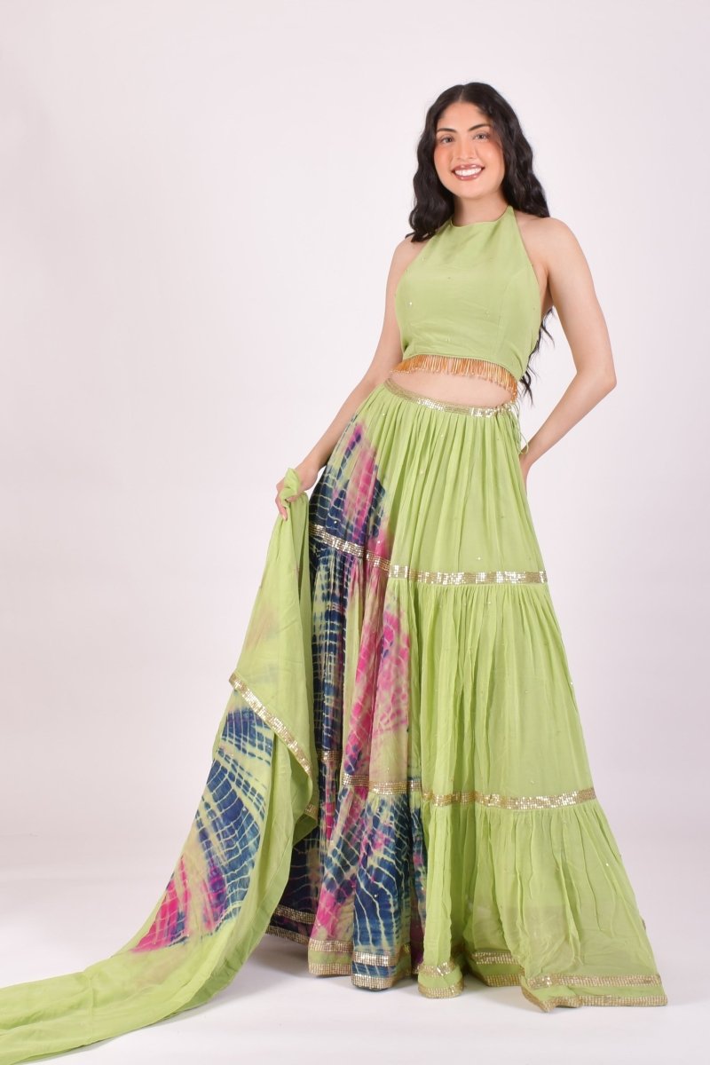 A woman modeling a light green traditional outfit with a sleeveless crop top, tiered skirt, and a matching dupatta featuring a blue, pink, and green tie-dye pattern with gold borders, set against a plain light background.