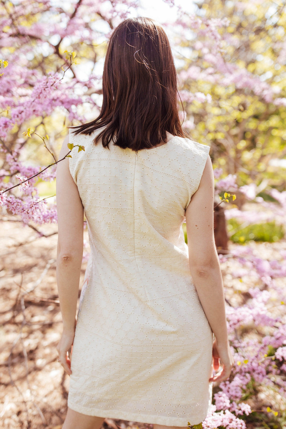 White cotton mini dress with all-over eyelet embroidery, sleeveless, shown from the back amidst pink blossoms.
