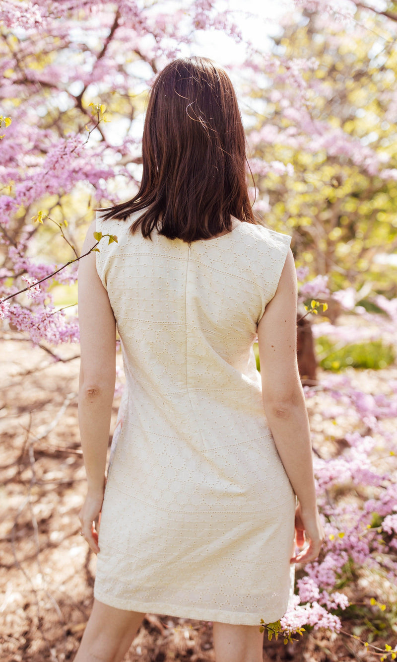 White cotton mini dress with all-over eyelet embroidery, sleeveless, shown from the back amidst pink blossoms.