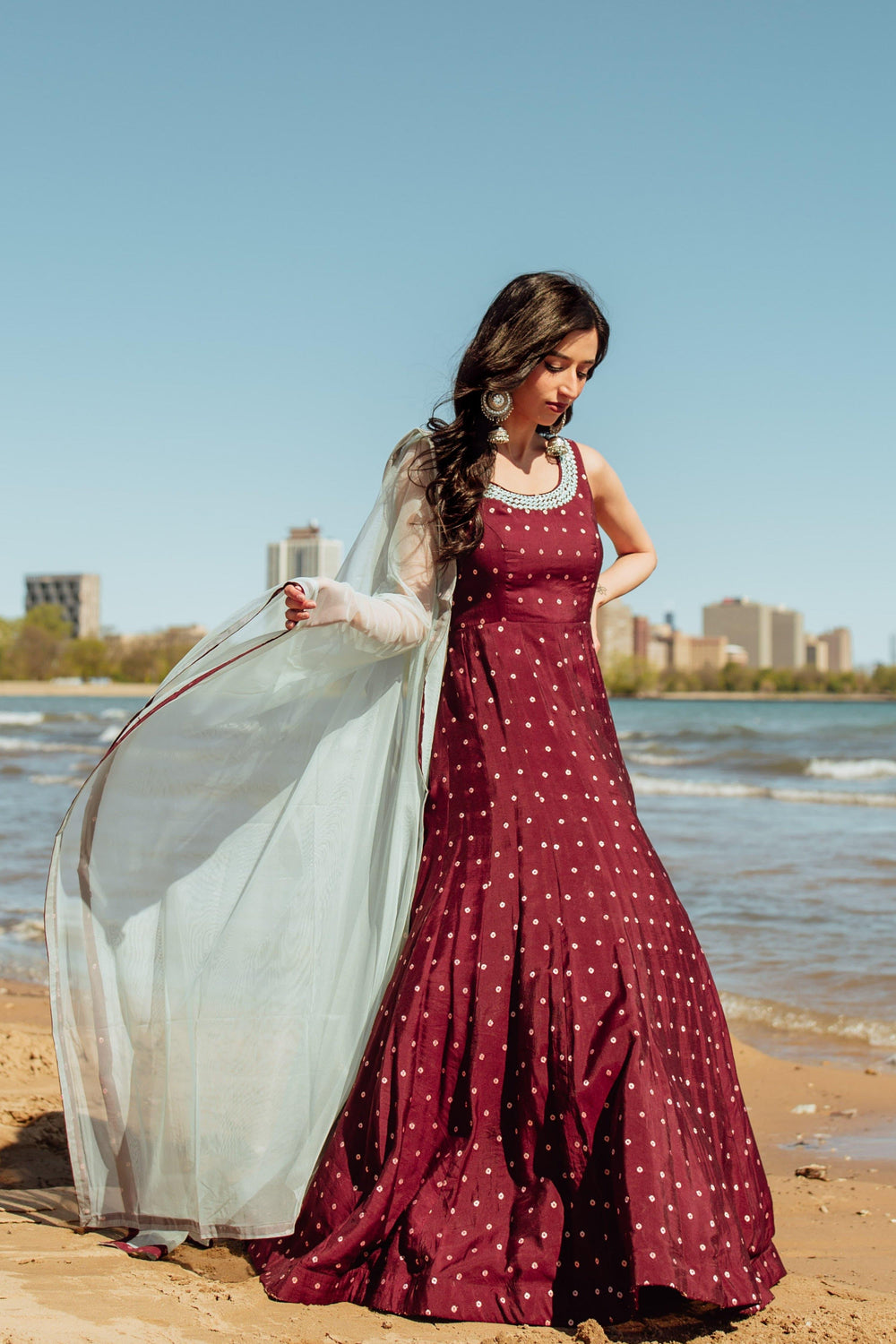 Wine silk A-line gown with resham embroidery, mirror work neckline, and flowing net drape on a beach.