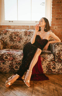 Wine velvet hand-embroidered off-shoulder long dress with a front slit, modeled sitting on a floral sofa.