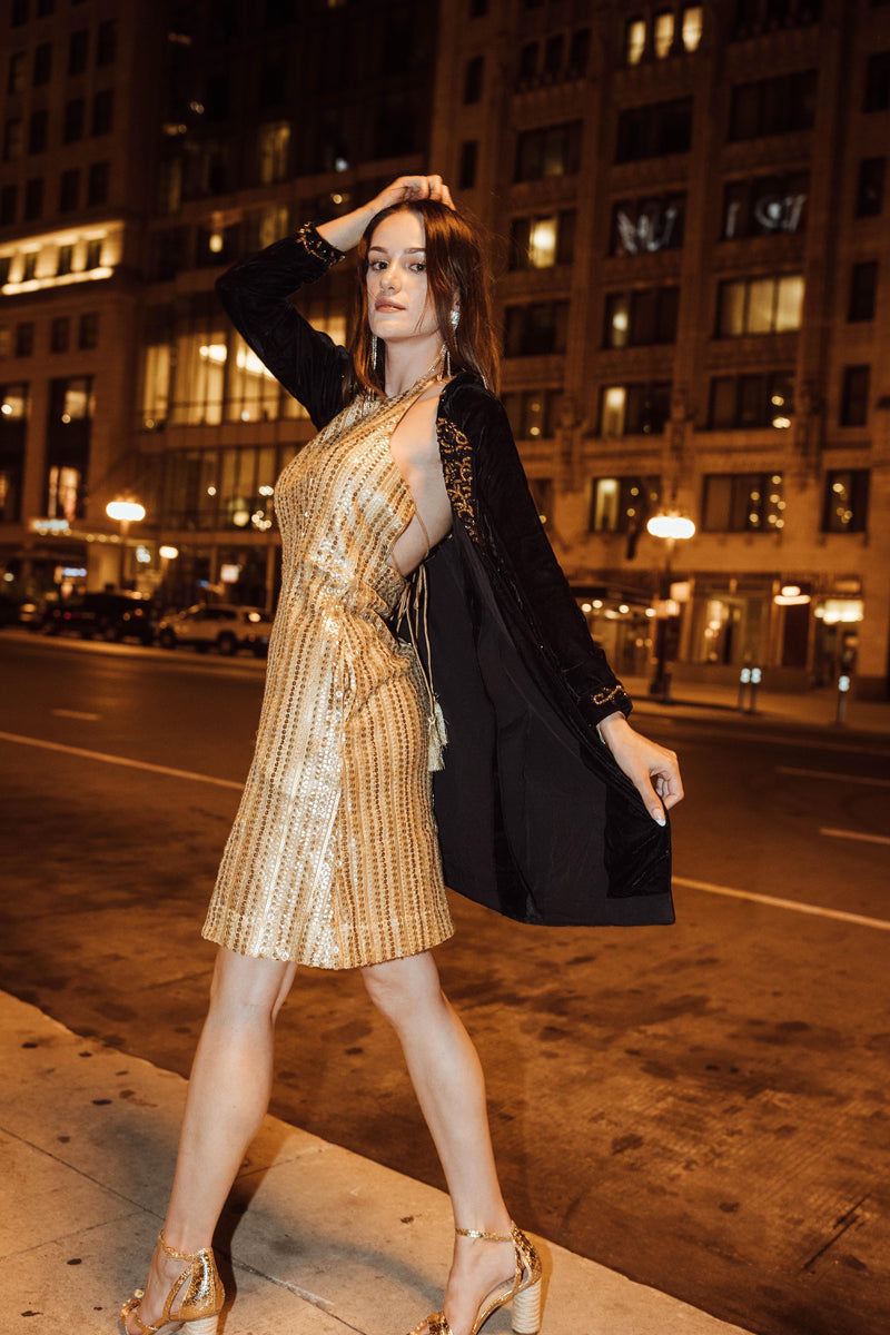 Model in black velvet blazer dress with gold embroidery over a sequined golden dress, posing on a city street at night.