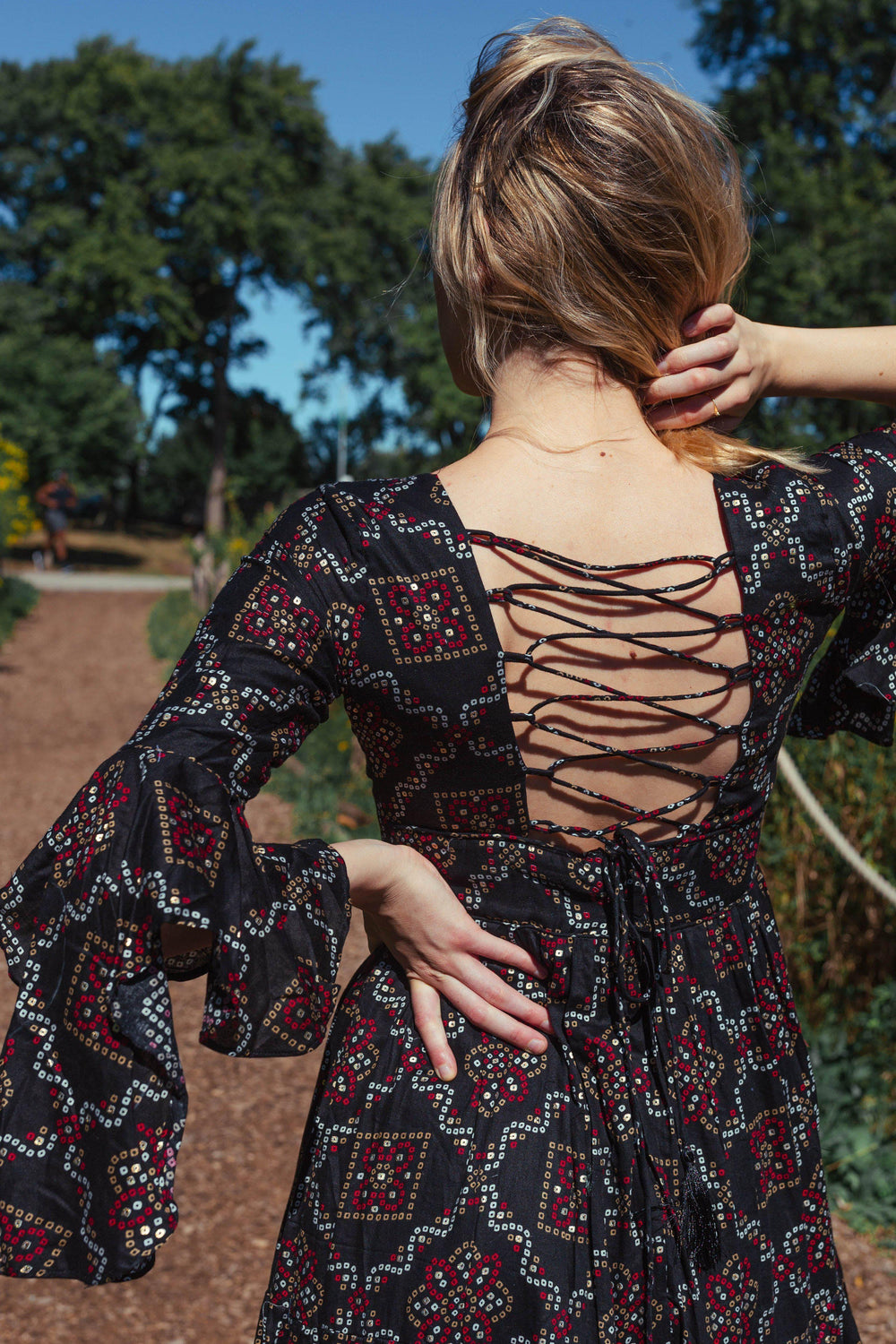Woman in black boho dress with red/gold print, lace-up back, and bell sleeves.