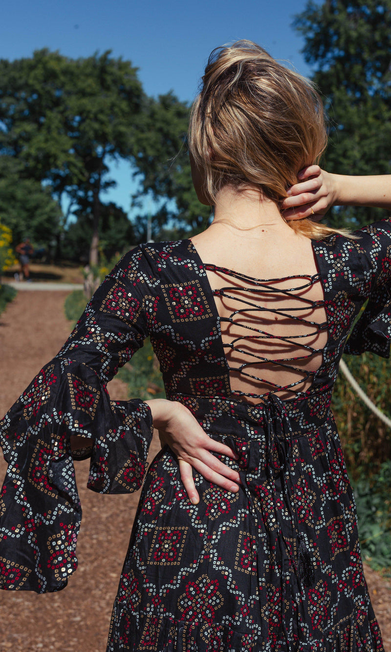 Woman in black boho dress with red/gold print, lace-up back, and bell sleeves.