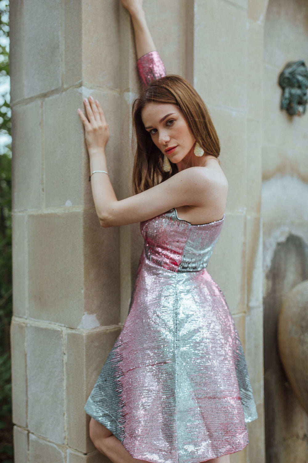 Model in a vibrant multi-color sequin one-shoulder mini dress, leaning against a stone structure.