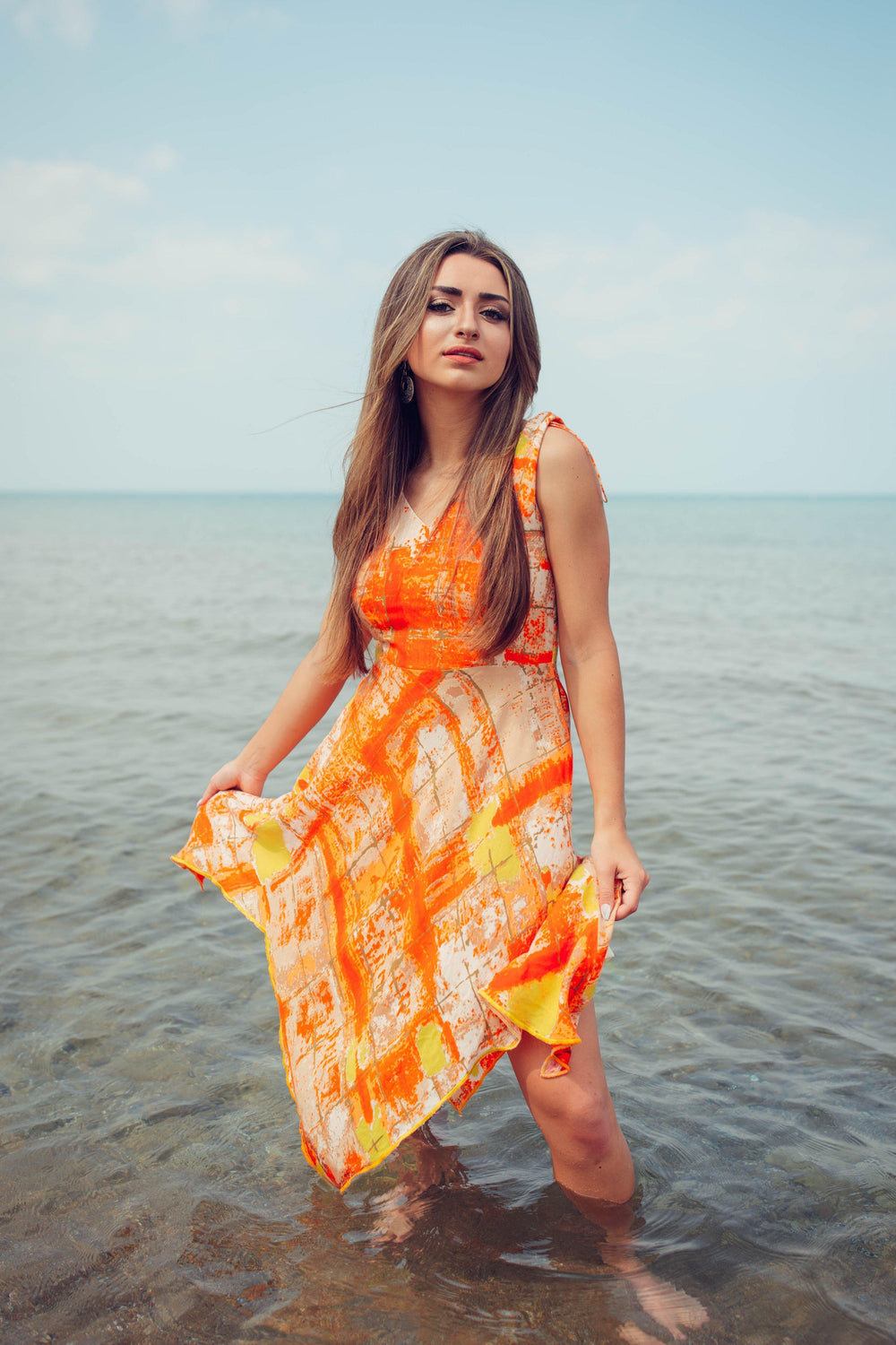 Woman in a vibrant orange tie-dye viscose rayon high-low dress, standing in shallow ocean water.