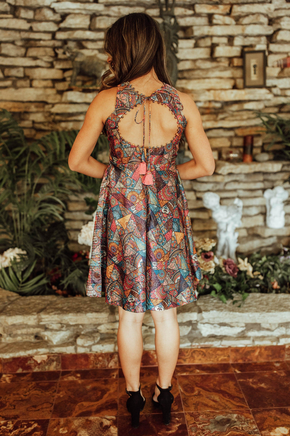 Back view of a woman in a sleeveless, mini cotton-silk dress with a colorful geometric-print skirt and an open back with tassel ties.