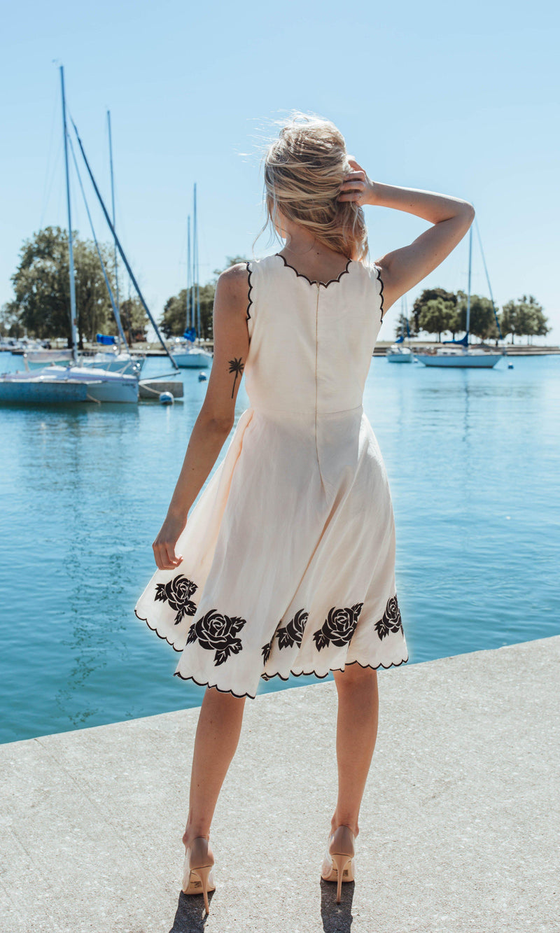 White modal silk mini dress with black embroidered roses on the scalloped hem, worn by a woman outdoors.